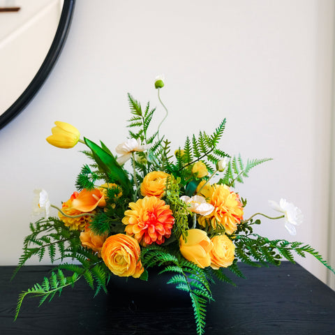 A vibrant artificial floral arrangement featuring yellow tulips, orange roses, and green ferns, displayed on a dark wooden table in front of a white wall with a round mirror.