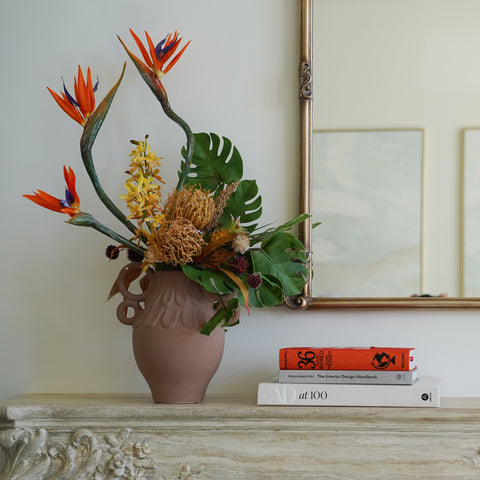 A terracotta-colored ceramic vase filled with a vibrant tropical floral arrangement, styled on a carved stone mantelpiece next to a stack of books. A large gold-framed mirror hangs on the cream wall behind, reflecting a window and brick wall.