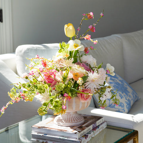 A vibrant floral arrangement in a white pedestal vase, styled on a glass coffee table next to a white sofa with blue patterned pillows.