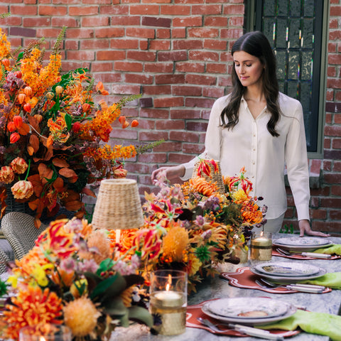 A woman in a cream shirt stands beside an outdoor dining table decorated with elaborate autumn floral arrangements in orange, burgundy, and gold tones, set against a red brick wall.
